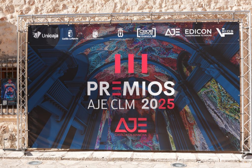 Lona promocional de los III Premios AJE Castilla-La Mancha 2025 colocada en un soporte metálico, con diseño colorido y logotipos de los patrocinadores sobre fondo de piedra.