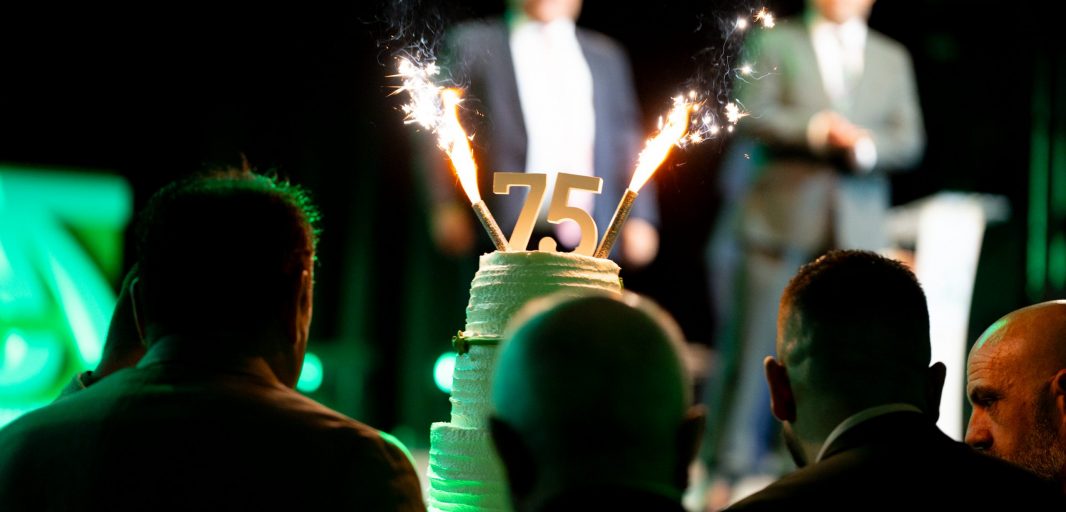 Tarta con el número 75 y bengalas en un evento de Sanehogar, con asistentes y ponentes en el escenario al fondo.