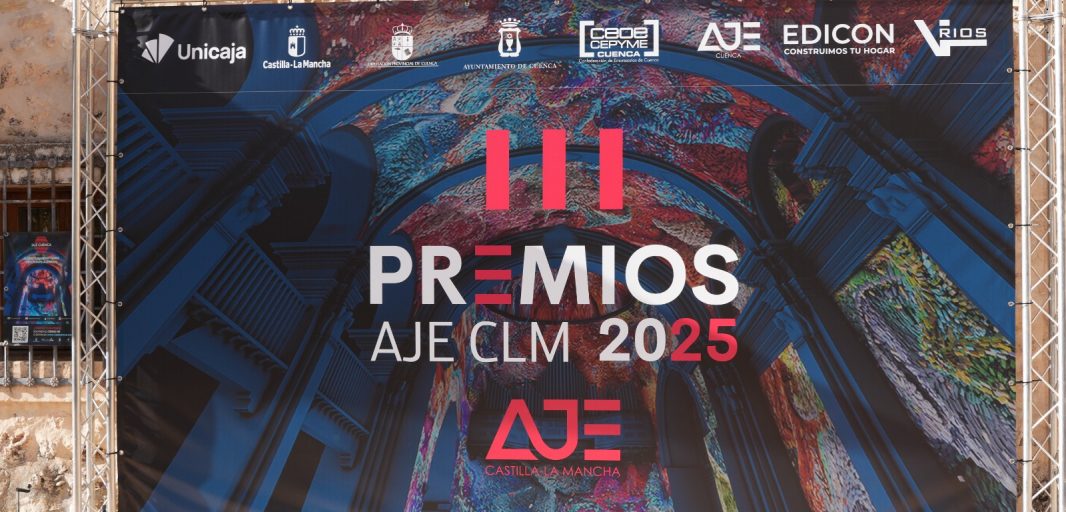 Lona promocional de los III Premios AJE Castilla-La Mancha 2025 colocada en un soporte metálico, con diseño colorido y logotipos de los patrocinadores sobre fondo de piedra.
